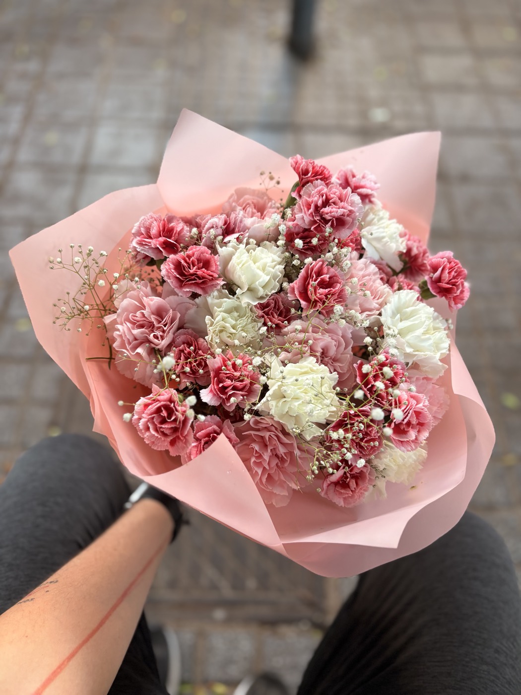 A bouquet of pink and white carnations with gypsophila, wrapped in pale pink paper and held on a lap on a tiled pavement.