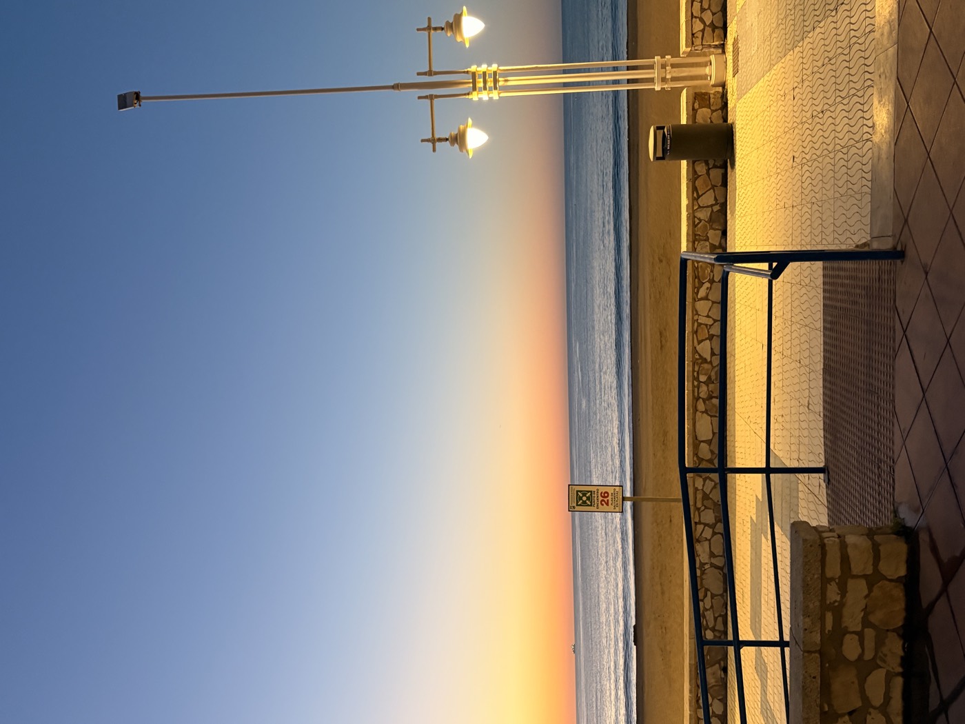 The seafront at dusk, a lamp post lit above an empty promenade with the sea and a long stretch of sand beyond.