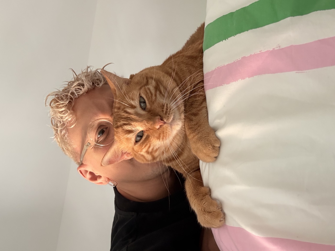 Igor lying cheek to cheek with Paco, a ginger cat, both looking at the camera over a pink and green striped pillow.