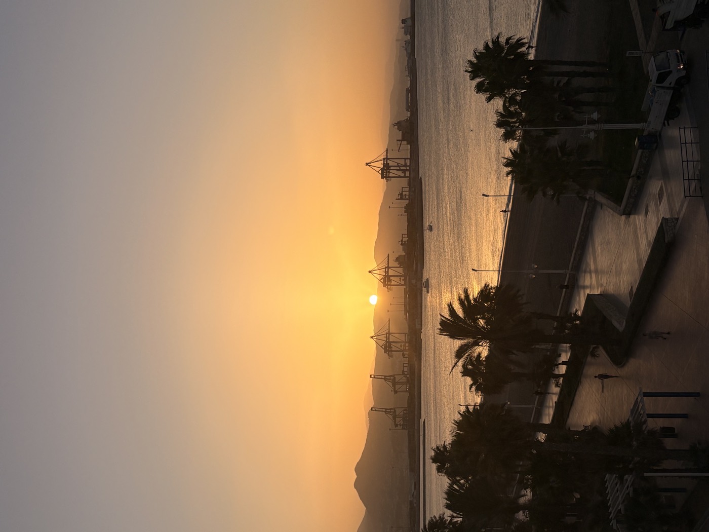 The sun dropping behind the hills over Málaga port, with cranes on the far quay and palm trees along the promenade below.
