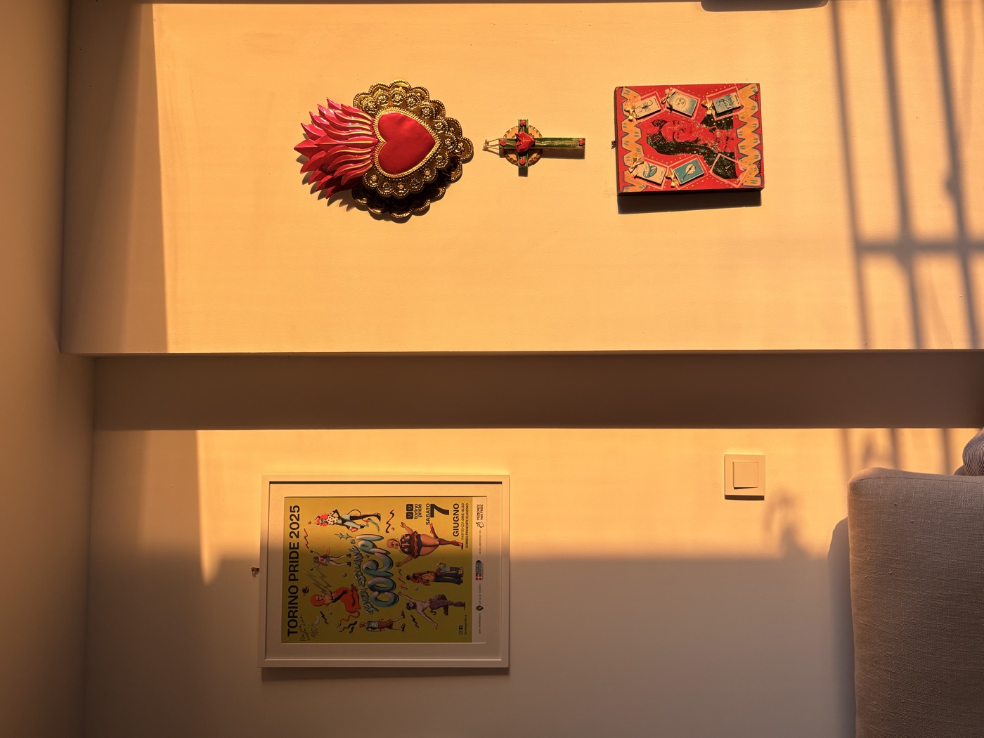 Late afternoon sun across a living room wall, with a framed Torino Pride 2025 poster on one side and a Mexican sacred heart, cross and painted box on the other.
