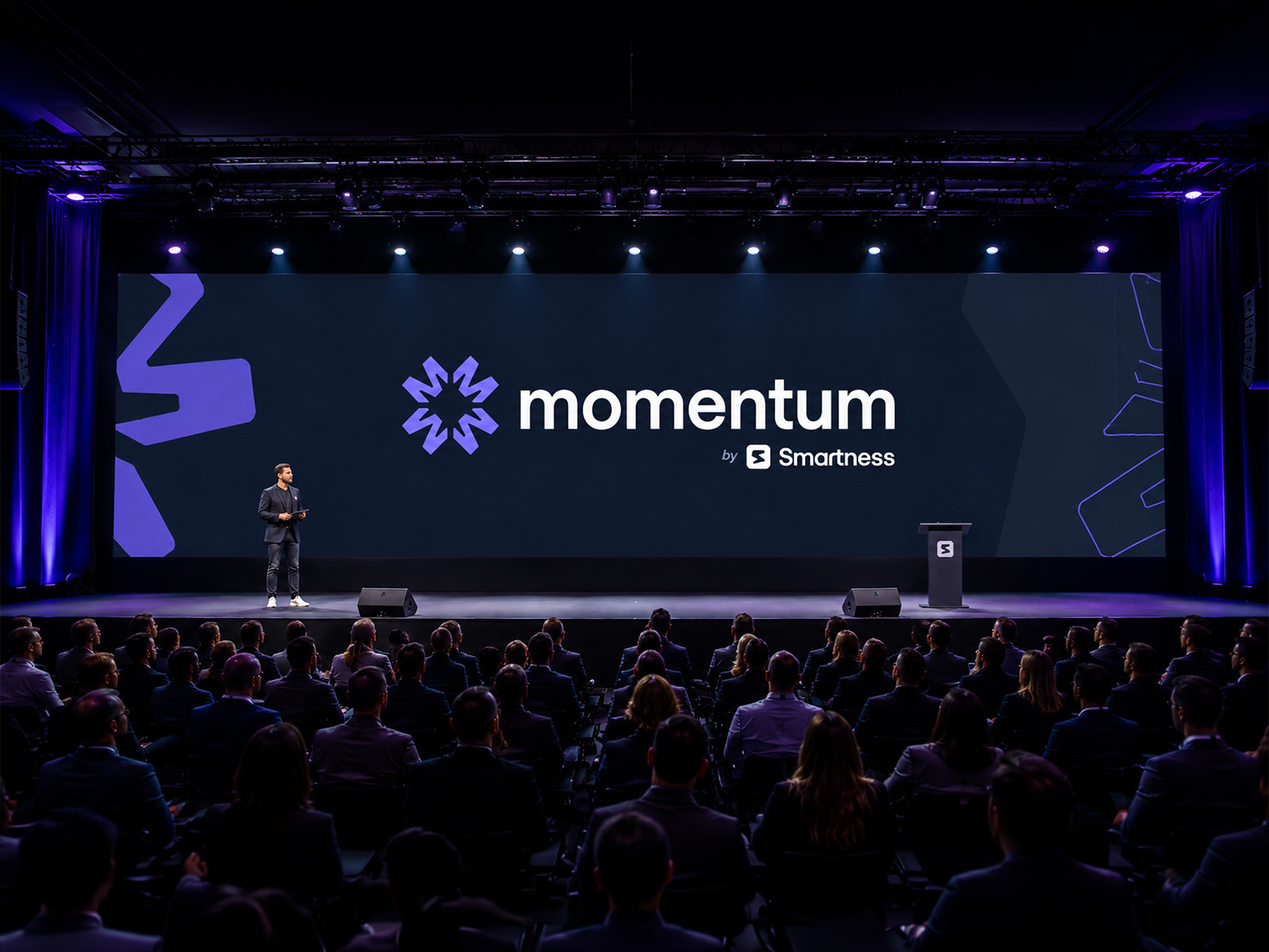The Momentum keynote stage, a speaker alone in front of a wide dark screen carrying the radial mark and the momentum wordmark, with a full auditorium seated in front of it.